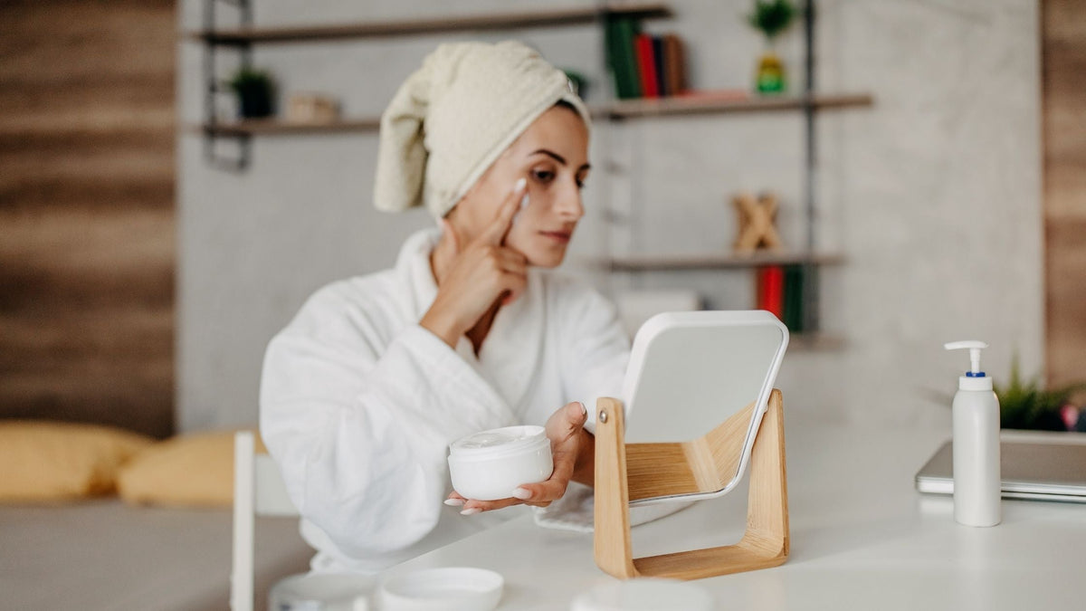 Woman in robe and towel applying skincare cream in front of mirror