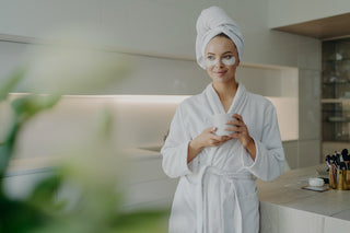 woman in bathrobe and towel on head drinking tea while doing skin care procedures at home