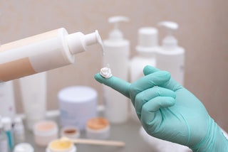 Hand squeezing a drop of cream from a bottle onto a hand in a glove on the background of cosmetic products, close up.